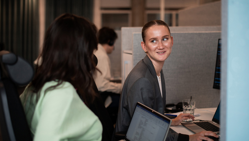Kvinnor samtalar vid skrivbord på kontor, en ler mot kameran.