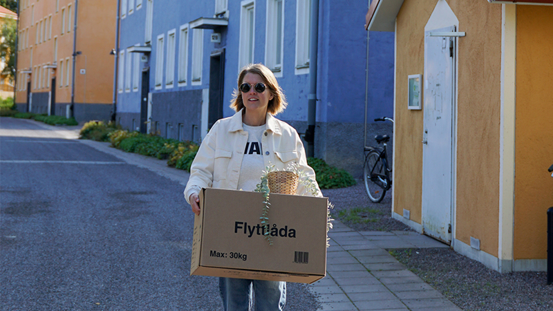 Person bär flyttlåda med texten "Flyttlåda" på gata mellan bostadshus.